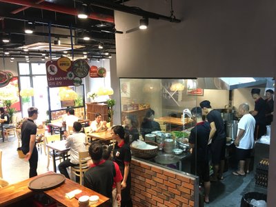Phòng ăn của Phở Thìn Hạ Long có thực khách ngồi bàn gỗ, phía sau là bếp quây gạch đỏ và kính.